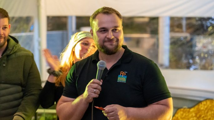 Mann mit Mikrofon lächelt in einer Veranstaltung, Publikum im Hintergrund.