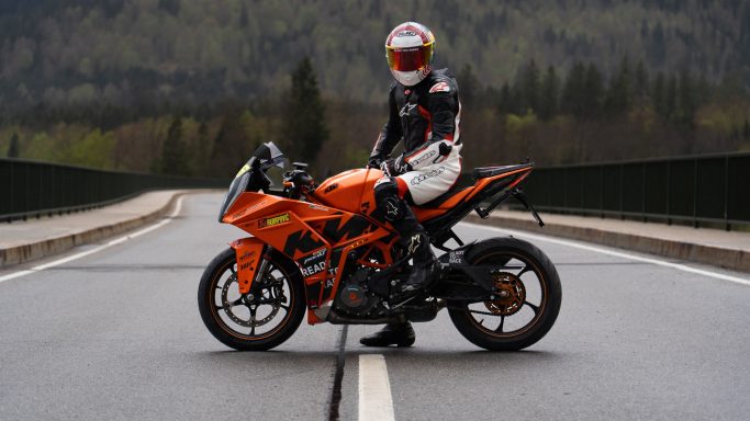 Ein Fahrer in Rennmontur steht auf einer orangen Sportmotorrad auf einer Straße.
