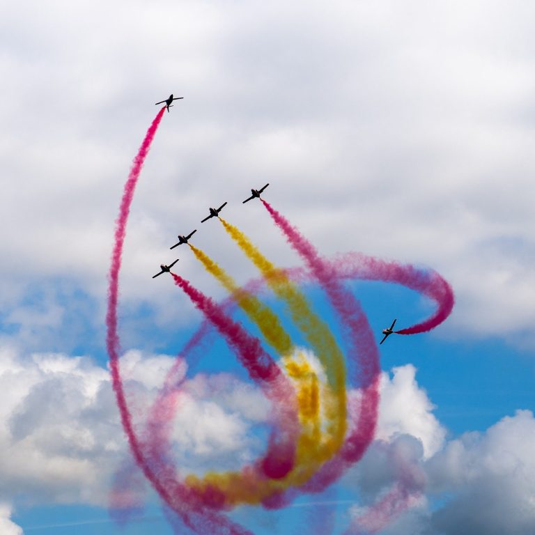 Fünf Flugzeuge bilden spiralförmige Rauchspuren in Pink und Gelb am Himmel.