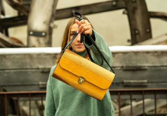 Eine Person zeigt eine gelbe Handtasche vor industrieller Kulisse.