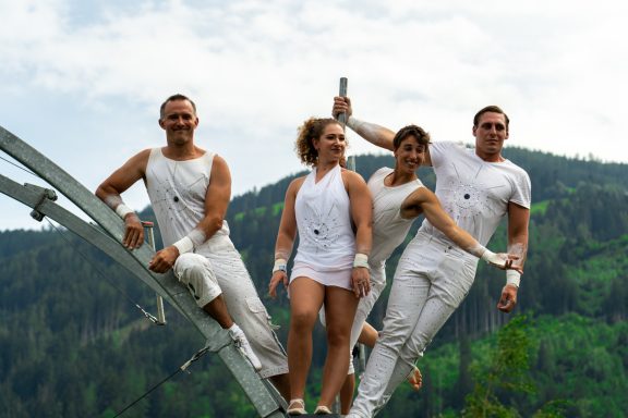 Vier Personen in weißen Outfits posieren auf einer Metallstruktur vor einer grünen Berglandschaft.