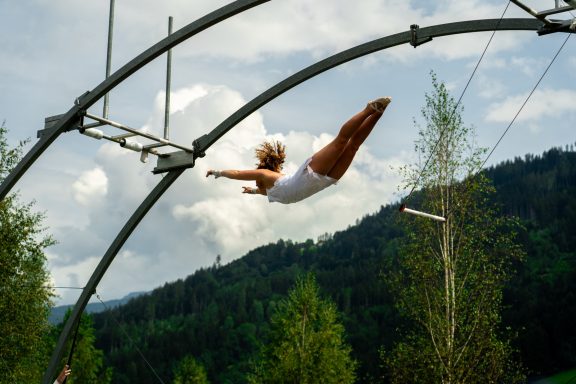 Frau springt von einem Trapez in die Luft, umgeben von Bäumen und Bergen.