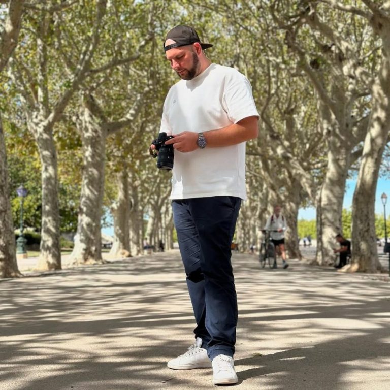 Mann in T-Shirt und Jeans steht in einer Baumallee und schaut auf seine Kamera