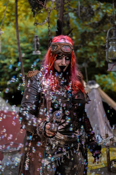 Person mit bunten Haaren und Kostüm, umgeben von Seifenblasen in einem Wald.