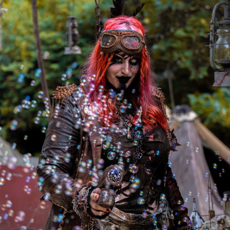 Person mit bunten Haaren und Steampunk-Kleidung, umgeben von Blasen im Freien.