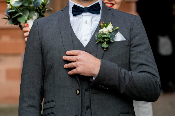 Mann in eleganter Hochzeitskleidung mit Fliege, Hand auf der Brust, Frau im Hintergrund.