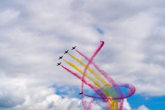 Sechs Flugzeuge fliegen in Formation mit bunten Rauchspuren am Himmel.