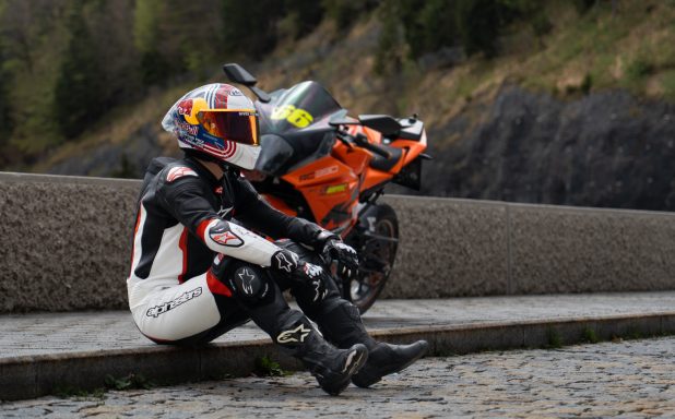 Motorradfahrer in Schutzkleidung sitzt neben einem orangefarbenen Motorrad.